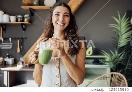 Attractive curly-haired girl in white top smiles and drinks green smoothie from transparent glass Attractive curly-haired girl in white top smiles and drinks green smoothie from transparent glass 123915950