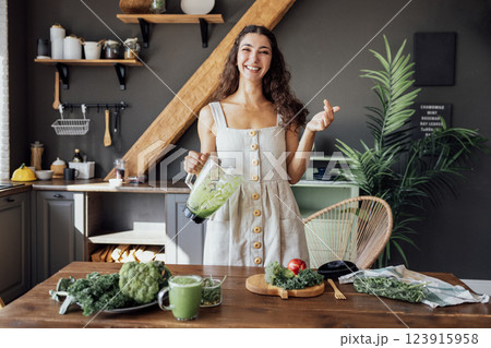A happy curly-haired girl in a linen sundress is preparing a healthy breakfast. A young smiling woman holds a blender with a green smoothie. A happy curly-haired girl in a linen sundress is preparing a healthy breakfast. A young smiling woman holds a blender with a green smoothie. 123915958