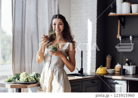 An attractive smiling girl is eating a salad of sprouted greens in the kitchen. A charming curly-haired brunette woman in a linen sundress holds a transparent bowl with micro greenery. An attractive smiling girl is eating a salad of sprouted greens in the kitchen. A charming curly-haired brunette woman in a linen sundress holds a transparent bowl with micro greenery. 123915960