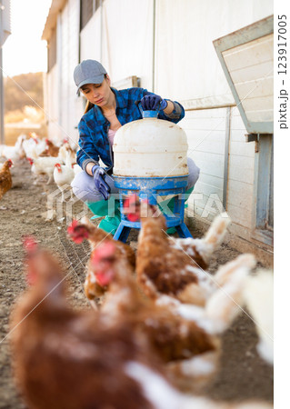 Female farmer holding legged bird drinker while chicken walking on backyard 123917005