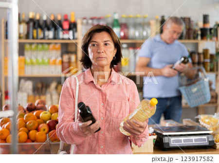Mature woman buyer looks thoughtfully at bottles of vegetable oil, man client in background Mature woman buyer looks thoughtfully at bottles of vegetable oil, man client in background 123917582
