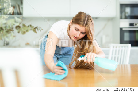 Attentive European teen girl in casual clothes spraying detergent on microfiber rag and wiping countertop in cozy cuisine Attentive European teen girl in casual clothes spraying detergent on microfiber rag and wiping countertop in cozy cuisine 123918408
