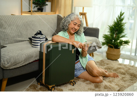 Cheerful mature woman sitting on the floor next to her suitcase and using smartphone Cheerful mature woman sitting on the floor next to her suitcase and using smartphone 123920860
