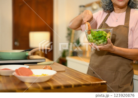 Cropped shot mature woman mixes fresh green salad in a glass bowl while standing in a kitchen 123920876