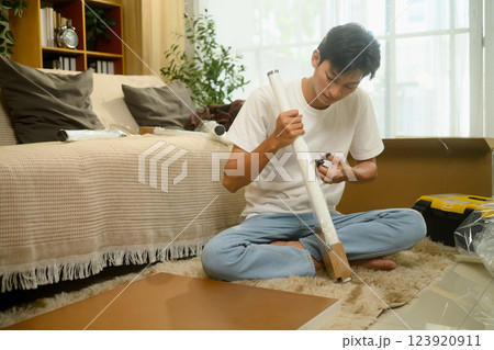 Young asian man unpacking a table leg preparing to assemble a new piece of furniture in his living room Young asian man unpacking a table leg preparing to assemble a new piece of furniture in his living room 123920911