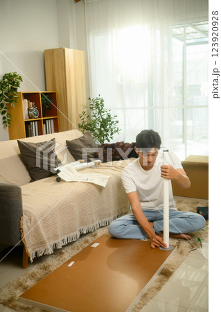 Young asian man sitting on soft rug assembling a table and following assembly instructions 123920928