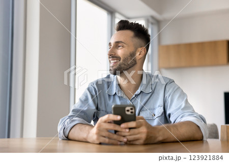 Happy Latin man looking away holding smartphone seated at table Happy Latin man looking away holding smartphone seated at table 123921884