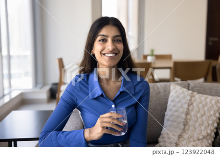 Indian woman holding glass of still water seated on sofa Indian woman holding glass of still water seated on sofa 123922048