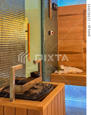 Heater with hot stones in sauna. Modern sauna interior with a wooden heater, walls, glass door, warm lighting. Relaxing wellness space with mosaic tiles and steam stones Heater with hot stones in sauna. Modern sauna interior with a wooden heater, walls, glass door, warm lighting. Relaxing wellness space with mosaic tiles and steam stones 123922643