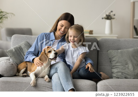 Lovely girl stroking dog seated on sofa with loving mother 123922655
