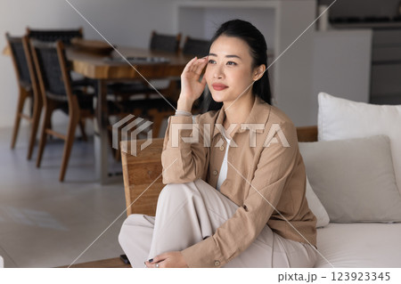 Positive dreamy young Asian woman sitting on sofa at home 123923345