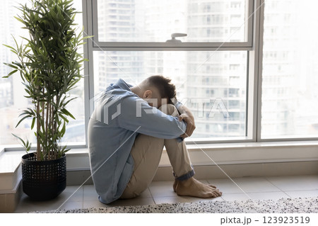 Deeply distressed or overwhelmed man sits on floor near window 123923519