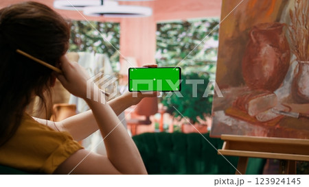Female artist holding device with green screen in art and craft atelier, admiring rustic artwork painted with watercolor paint. Woman preparing masterpiece for art gallery exhibition. Camera B. Female artist holding device with green screen in art and craft atelier, admiring rustic artwork painted with watercolor paint. Woman preparing masterpiece for art gallery exhibition. Camera B. 123924145