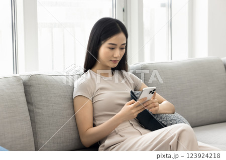 Serious Asian woman sitting on sofa hold smartphone Serious Asian woman sitting on sofa hold smartphone 123924218