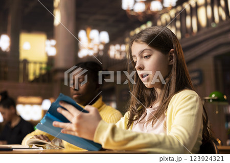 Two diverse schoolgirls in a library reading books for a school project, concentrating on curriculum based knowledge and literary exploration. Young scholars focus on mandatory lectures. Two diverse schoolgirls in a library reading books for a school project, concentrating on curriculum based knowledge and literary exploration. Young scholars focus on mandatory lectures. 123924321