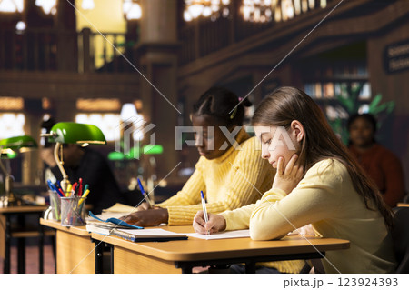 Two diverse teenagers focused on writing an essay in a public library, completing their school assignment with pens and notebooks. Girls showcasing concentration and academic development. Two diverse teenagers focused on writing an essay in a public library, completing their school assignment with pens and notebooks. Girls showcasing concentration and academic development. 123924393