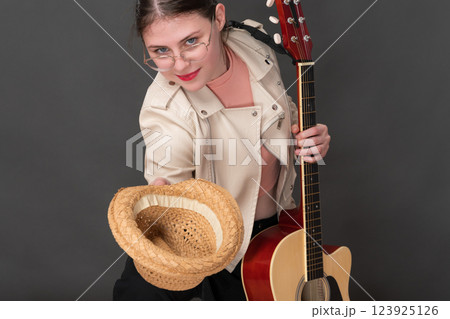 Young female guitarist with guitar in one hand and straw hat in other hand stretched forward. Creativity street musician asks for donation for playing guitar. Playful woman looking at camera 123925126