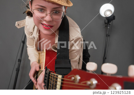 Cheerful female guitarist with glasses and grin on face is holding guitar and pointing it at camera. Playful expression musician is having fun, enjoying playing guitar during performance on stage Cheerful female guitarist with glasses and grin on face is holding guitar and pointing it at camera. Playful expression musician is having fun, enjoying playing guitar during performance on stage 123925128