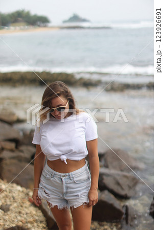 A woman in a white crop top and shorts stands on a rock by water 123926691