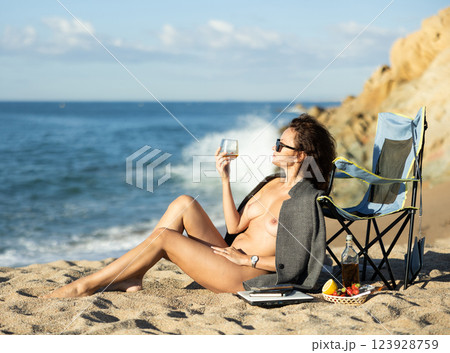 Curvy brown haired woman in suit jacket and sunglasses drinking whiskey on beach 123928759
