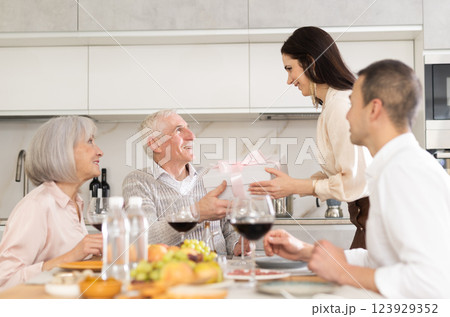 Young woman giving present to old man while sitting around kitchen table 123929352