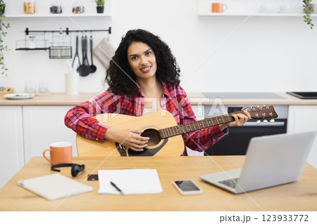 Young adult woman practicing guitar playing in home kitchen wearing plaid shirt, smiling. Relaxed and creative atmosphere with notebook and coffee mug on wooden table, expressing hobbies. 123933772