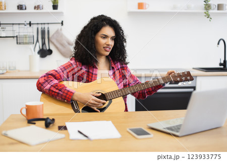 Young adult woman with curly hair playing guitar, watching online tutorial on laptop in kitchen. Home learning environment, creative self-improvement, indoor activity emphasizing personal development. Young adult woman with curly hair playing guitar, watching online tutorial on laptop in kitchen. Home learning environment, creative self-improvement, indoor activity emphasizing personal development. 123933775