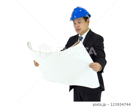 A man in a business suit wearing a blue hard hat thoroughly inspects large architectural plans. He stands focused, preparing for a construction or renovation project. 123934744