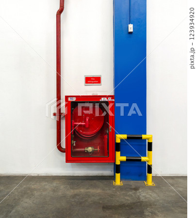 A fire hose reel is mounted on a white wall, accompanied by a bright red sign. A yellow and black safety barrier line the area, ensuring visibility and safety. A fire hose reel is mounted on a white wall, accompanied by a bright red sign. A yellow and black safety barrier line the area, ensuring visibility and safety. 123934920