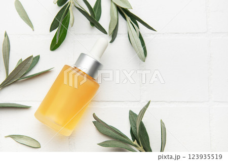 Natural cosmetic serum bottle with dropper surrounded by olive tree leaves on white tile background 123935519