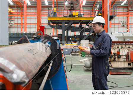 A man in a hardhat and mechanic coverall dust protection suit operates industrial equipment inside a busy factory setting, skillfully managing the machinery. 123935573