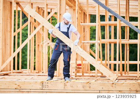 Carpenter constructing wooden-framed house. Man holding large joist and dressed in work clothes and helmet. Concept of modern and environmentally-friendly construction. Carpenter constructing wooden-framed house. Man holding large joist and dressed in work clothes and helmet. Concept of modern and environmentally-friendly construction. 123935925