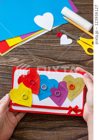 Child holding Greeting card with a hearts Valentine's Day, day mother or birthday present on wooden table. Childrens art project crafts for children. Craft for kids. 123936127