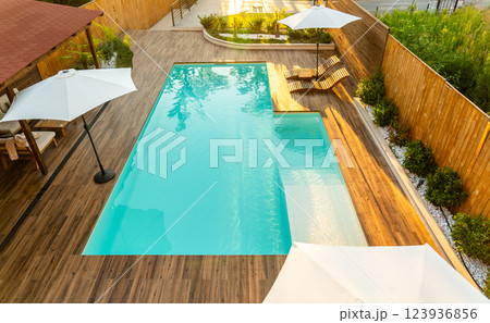 Elegant backyard with turquoise swimming pool, wooden deck, umbrellas, and lounge chairs in a sunny setting 123936856