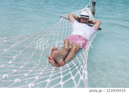 A young man in summer clothes and a hat is lying in a hammock in clear sea water A young man in summer clothes and a hat is lying in a hammock in clear sea water 123939535