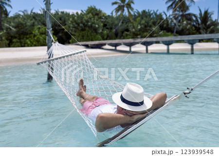 A young man in summer clothes and a hat is lying in a hammock in clear sea water A young man in summer clothes and a hat is lying in a hammock in clear sea water 123939548