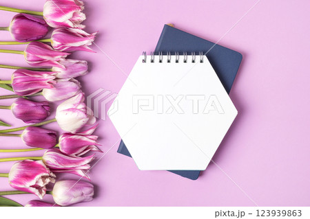 Open paper notebook with empty page with purple and white tulips on purple background. Top view, flat lay, work space on purple background. Open paper notebook with empty page with purple and white tulips on purple background. Top view, flat lay, work space on purple background. 123939863