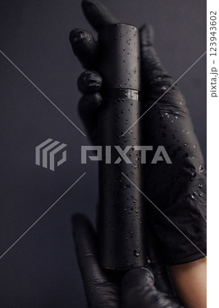 A mockup of a black tube for cream with drops of water. Close-up of the beautician hands in black gloves holding a moisturizer on a dark isolated background. 123943602