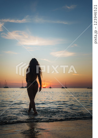 A serene beach scene at sunset in Phuket or Koh Phi Phi, Thailand, shows a woman entering calm waters with silhouetted sailboats on the horizon. A serene beach scene at sunset in Phuket or Koh Phi Phi, Thailand, shows a woman entering calm waters with silhouetted sailboats on the horizon. 123946521
