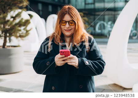 Happy woman chatting on phone and relaxing outdoors. Urban, people concept. Smiling 30s redhead woman text messaging on smart phone at sunny day on the city street, spring time Happy woman chatting on phone and relaxing outdoors. Urban, people concept. Smiling 30s redhead woman text messaging on smart phone at sunny day on the city street, spring time 123948538