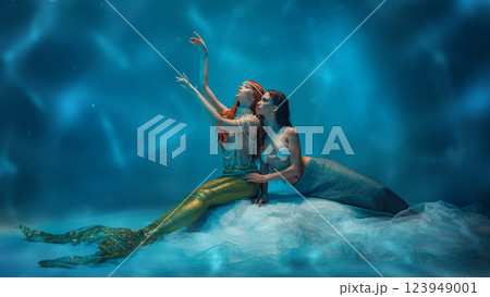 Mystical mermaid extending arms towards glowing light as another leans into her, bathed in deep blue reflections. Mystical mermaid extending arms towards glowing light as another leans into her, bathed in deep blue reflections. 123949001