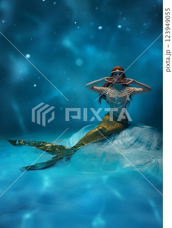 Mystical underwater mermaid, redhaired woman with body covered in pearls, sitting deep in ocean against water-like wavy background. 123949055