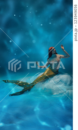 Graceful young redhaired woman, mermaid with golden tail floating in dreamy blue water, surrounded by soft light. 123949056