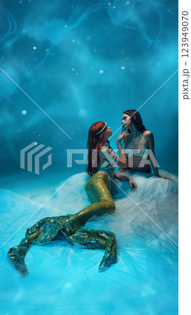 Two passionate young women, mermaids sitting deep in the ocean, executing love and tenderness against blue glowing background 123949070