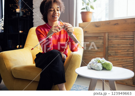 Young African smiles and knits at home in living room. Black girl in causal clothes is engaged in needlework Young African smiles and knits at home in living room. Black girl in causal clothes is engaged in needlework 123949663