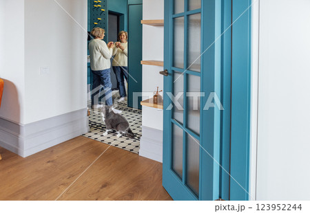 An elderly woman stands at the mirror and paints her lips in the hallway of the apartment. Mature lady in casual clothes applying decorative cosmetics. An elderly woman stands at the mirror and paints her lips in the hallway of the apartment. Mature lady in casual clothes applying decorative cosmetics. 123952244