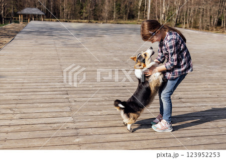 A little girl hugs her corgi puppy on the street. A cute kid in casual clothes is laughing and playing with her dog on a wooden platform. A little girl hugs her corgi puppy on the street. A cute kid in casual clothes is laughing and playing with her dog on a wooden platform. 123952253