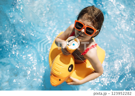 Little girl in sunglasses and swimsuit drinks cocktail in pool. Cute girl enjoys milkshake and swim in water. Little girl in sunglasses and swimsuit drinks cocktail in pool. Cute girl enjoys milkshake and swim in water. 123952282