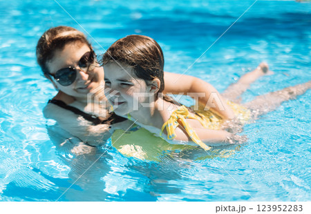A young woman in sunglasses teaches her little daughter to swim. An attractive mom and her cute girl are swimming in the pool. 123952283