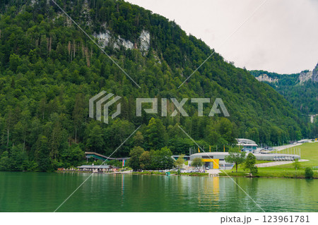 Vintage wooden boathouses line the calm, green waters, surrounded by dense trees and towering mountains under a cloudy sky, creating a tranquil atmosphere in nature. Konigssee lake Bavaria German Alps 123961781